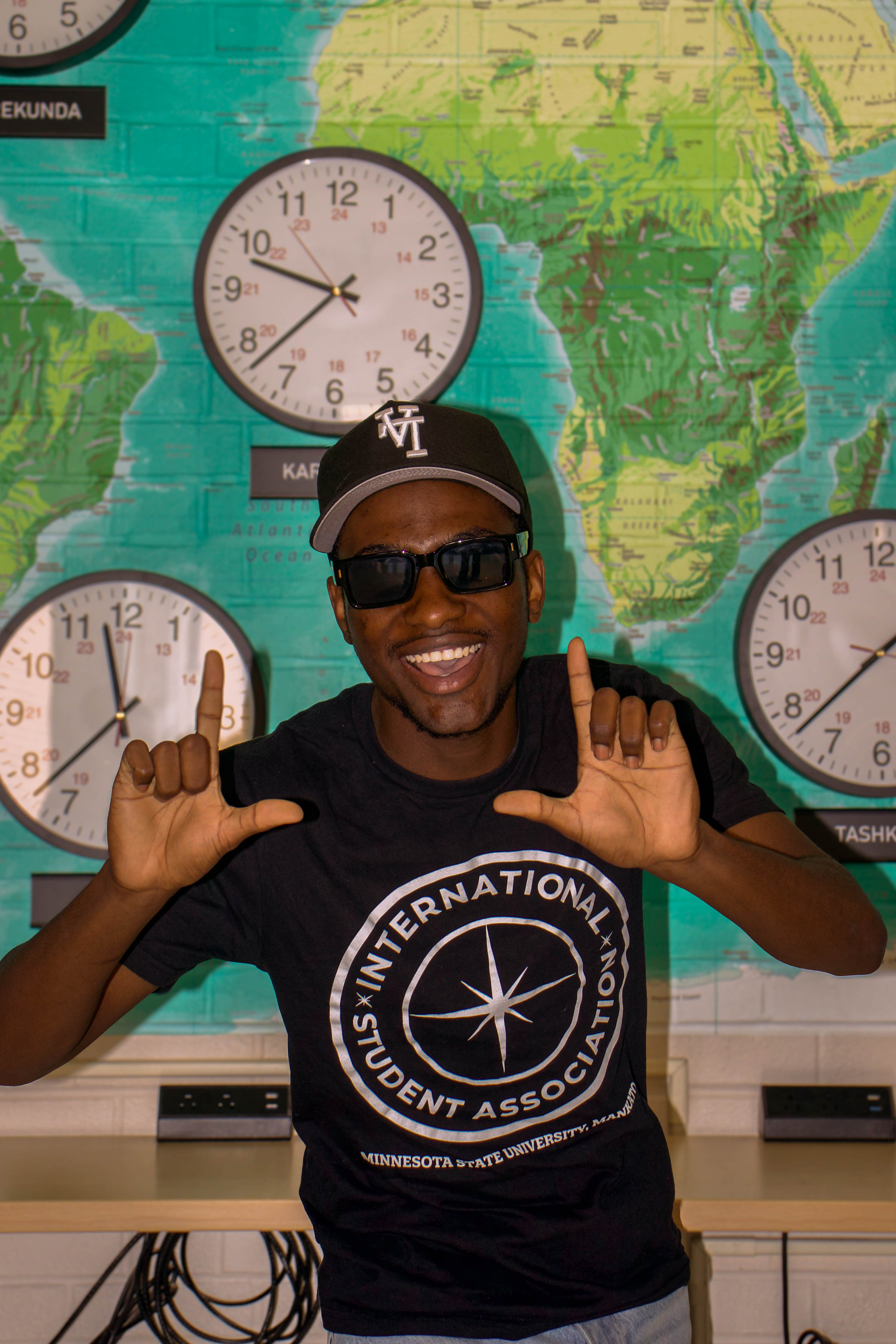 Portrait of Farai smiling widely with sunglasses and a baseball cap, wearing a black International Student Association t-shirt and gesturing with his hands in front of a teal world map wall and clocks.
