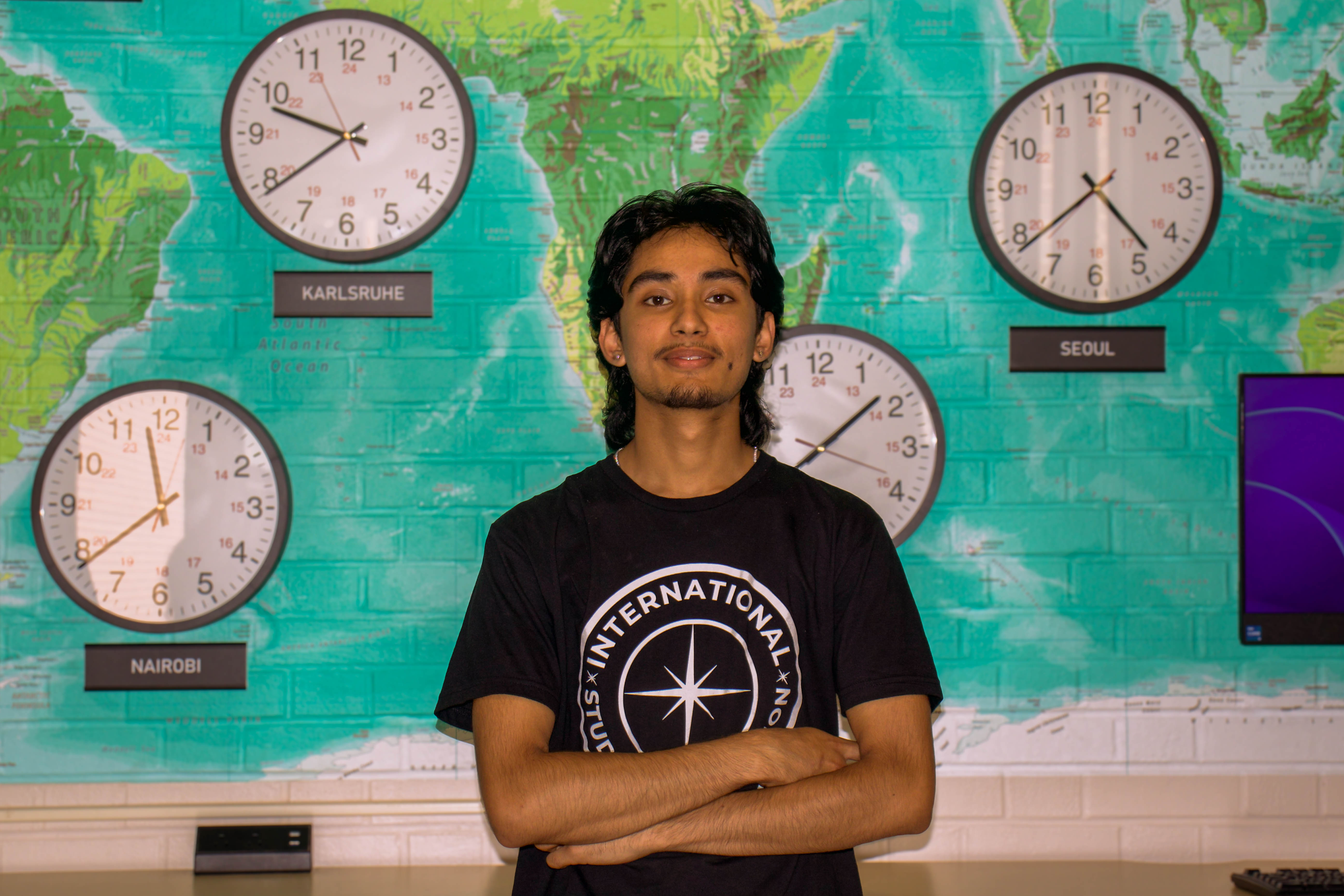 Portrait of GC standing with arms crossed, wearing a black International Student Association t-shirt in front of a teal world map wall and clocks.