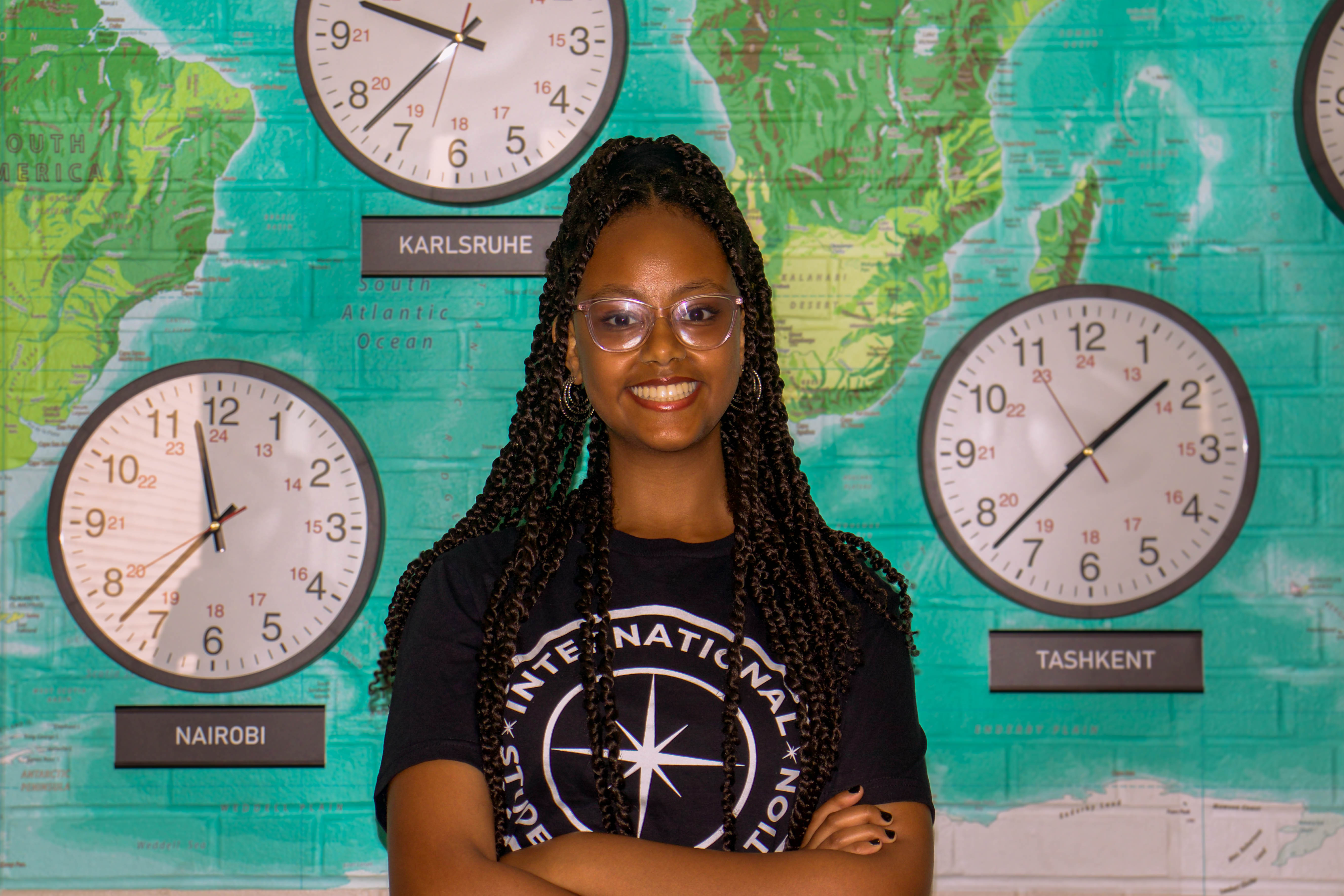 Portrait of Gelila smiling with braided hair, glasses, and a black International Student Association t-shirt, standing with arms crossed against a teal world map wall and clocks.