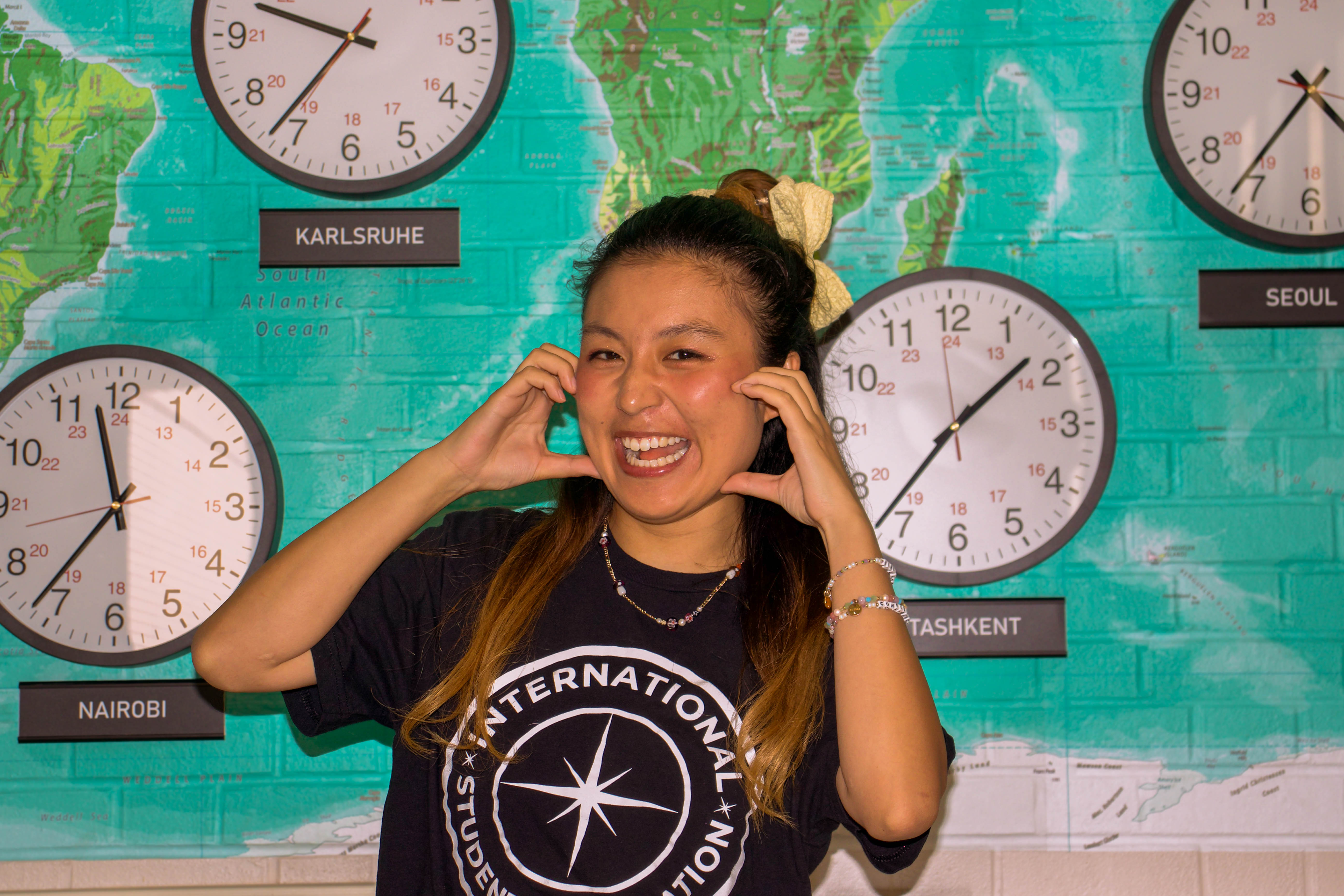 Portrait of Riko smiling joyfully with her hands framed around her cheeks, wearing a hair bow and a black International Student Association t-shirt against a teal world map wall and clocks.